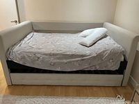 Side view of pale pink fabric trundle bed with memory foam mattress, white V-shaped pillow on top, showing padded arms and wooden frame beneath mattress.