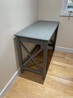 Blue-grey painted wooden desk with X-braced sides against wall on wooden floor.