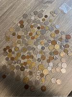 Wide view of approximately 100 assorted foreign coins of various sizes and colors spread on wooden floor.