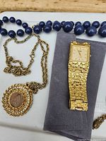 Photo showing blue bead necklace, gold-tone chain with coin and rhinestone pendant, and gold-tone textured wristwatch.