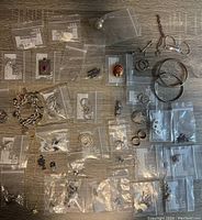 Overhead photo showing the full assortment of jewelry pieces laid out on a surface, many in small clear plastic bags. Includes bracelets, pendants, rings, earrings, and chains in silver and gold tones.