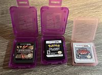 Three Pokemon Nintendo game cartridges in open plastic cases on wood surface: Japanese Pokemon Platinum DS cartridge in purple case, English Pokemon Black Version DS cartridge in purple case, Pokemon Rumble Blast 3DS cartridge in pink case.