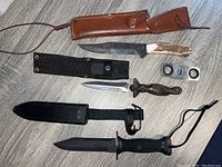 Four cutting tools arranged on floor including Bowie knife with leather sheath, black tactical knife and sheath, mermaid-handled dagger, cigar cutter