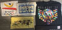 Four vintage T-shirts laid flat, showing front printed graphics: white Barcelona 1992 Olympics, cream and blue World Cup Soccer 1994, black World Cup USA 94, and yellow Crazy Shirt 1971 Hawaii.
