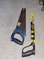 Photo showing two hand saws on a concrete floor. One has a blue and black handle with a rusty blade, the other has a yellow and black handle with a Stanley label.