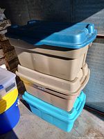 Three large plastic totes stacked; top two have beige bodies with blue and beige lids, the bottom one has a beige body with teal lid.