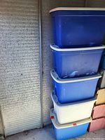 Five stackable plastic storage totes, four blue and one white, stacked vertically against a metal wall.