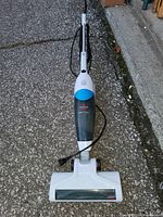 White Bissell stick vacuum cleaner with cord and cleaning brush head, photographed outdoors on a concrete surface.