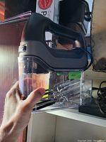 Side view of the black and silver hand blender held in hand, showing clear container with metal beaters inside, taken to show overall shape and attachments included.