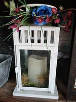 White wooden lantern with glass panels containing an artificial candle and moss, decorated on top with blue and pink artificial flowers and greenery.