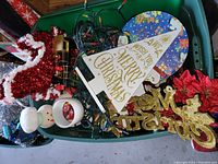 Photo of an open green plastic storage bin containing assorted Christmas decorations including a red and white tinsel stocking, string lights, gold glitter text signs, and a white triangular Christmas decoration.