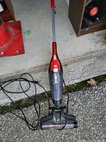 Photo showing the Dirt Devil stick vacuum placed on ground with power cord partially coiled, handle upright, clear dust container and brush head visible.
