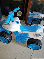 Side view of a white and blue kids quad bike riding toy featuring Frozen II graphics, four large wheels, a blue handlebar with character images, and a blue seat and footrest.