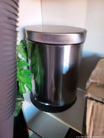 Photo of a round stainless steel garbage can with a lid sitting on a shelf beside some cardboard and plastic tubing.