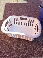 Image showing a white plastic laundry basket placed on asphalt outdoors. The basket has ventilation holes along the sides and handles on two opposite ends.