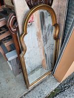 Photo showing the gold framed arched wall mirror leaning against a wall outdoors. The frame has a curved arch top with ornamental side detailing. Mirror glass surface is intact.