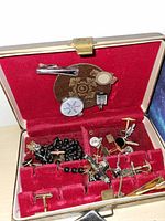 Open brown textured box with red velvet interior showing assorted small metal items including cufflinks, chain, beads, and pins inside.