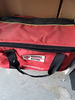 Closed red fabric zippered bag with black handles and label 'Roadside Assistance Winter Essential Kit'