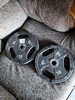 Two black metal 10 lb weight plates with grip holes and white numbering placed on a grey fabric surface.
