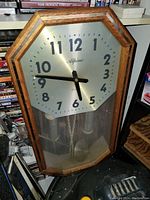 Wall clock with octagonal wooden frame, silver dial, Arabic numerals, black clock hands, and visible pendulum and weights inside.
