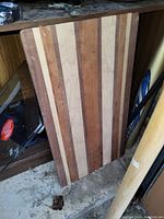 Photo of a rectangular wooden cutting board featuring alternating light and dark wood stripes. The cutting board leans against a wooden cabinet and shows some wear marks on the surface.