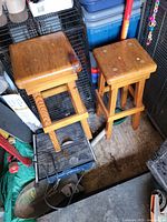 Two wooden bar stools with square seats, some wear on top surfaces, positioned side by side in a cluttered space.