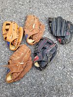 Five used baseball gloves of mixed brands and colors on asphalt surface