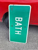 Green and white wooden sign with the word 'BATH' leaning against a red vehicle with visible wear and rust spots.