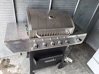 Closed view of the Nexgrill gas BBQ showing five burner control knobs, side burner, wheels and temperature gauge on stainless steel lid.