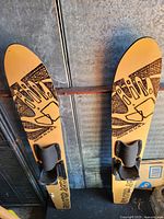 Pair of yellow Body Glove water skis with black binding straps, photographed hanging vertically against a metal wall