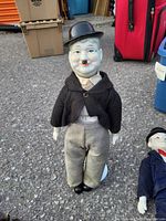 Front view of vintage porcelain Charlie Chaplin doll dressed with black hat, black jacket, gray pants, white shirt, standing on asphalt outdoors.