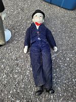 Photo of a vintage male doll dressed in a blue outfit with black shoes and hat lying on the ground outside.