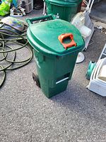 Photo of a green plastic garden compost bin with a hinged lid and orange handle, two black wheels for mobility, placed on asphalt.