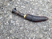 Photo of traditional kukri knife sheathed on asphalt surface showing dark leather sheath with stitching and belt loop.
