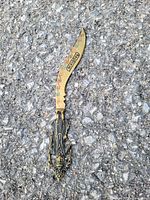 Photo of a curved dagger with 'PANAMA' etched on the blade lying on an asphalt surface. The handle features intricate gold and dark metal designs with a serrated blade section near the handle.