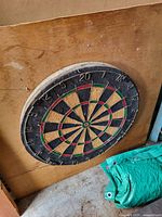 Dart board mounted on wooden board shown leaning against a wall with green tarp on floor.