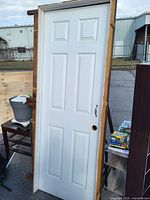 Front view of white six-panel door with frame and metal handle, leaning against a wooden structure outdoors.
