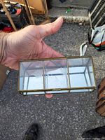 Small rectangular glass display case held in hand to show size and construction. Glass top, bottom and sides with brass-colored frame and three internal compartments.