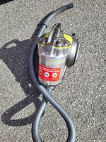 Hoover vacuum cleaner outside on pavement showing main unit with clear cyclone dust container, grey hose attached.