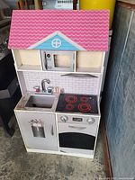 Front view photo of a wooden kids play kitchen set featuring a pink roof, sink with faucet, stove with four burners, oven with knobs, microwave, and lower cabinets.
