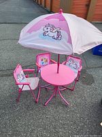 Pink kids table set with umbrella and four matching chairs featuring unicorn graphics, photographed outdoors on pavement