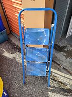 Photo of a blue metal step ladder with three steps showing paint wear and chipped paint on steps and frame.