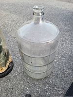 Clear large glass jug with round stopper viewed outdoors on pavement. Shows ribbed design around the body and stopper on top.