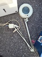 White adjustable magnifying lamp with clamp base and light, laying on asphalt surface next to other items.