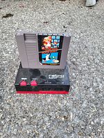 Photo showing a black and red Retro NES console with two red buttons on the front and a gray NES game cartridge labeled with Super Mario Bros. and Duck Hunt.