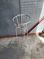 Full view of metal wire plant stand showing circular holder and tripod legs, placed on a concrete floor with metal wall background.