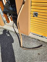 Single image of a traditional handheld scythe with a wooden handle and a curved metal blade placed upright on the ground outside a storage unit.
