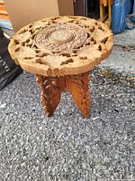 Blue-tinted photo of a small carved wooden round side table on an asphalt surface.