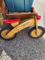 Photo of wooden Skuut balance bike with red seat and red handle grips, black tires, and yellow rims, resting on pavement.