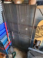 Black metal wire mesh small animal cage in storage area, showing front view with visible structure and hanging toys.